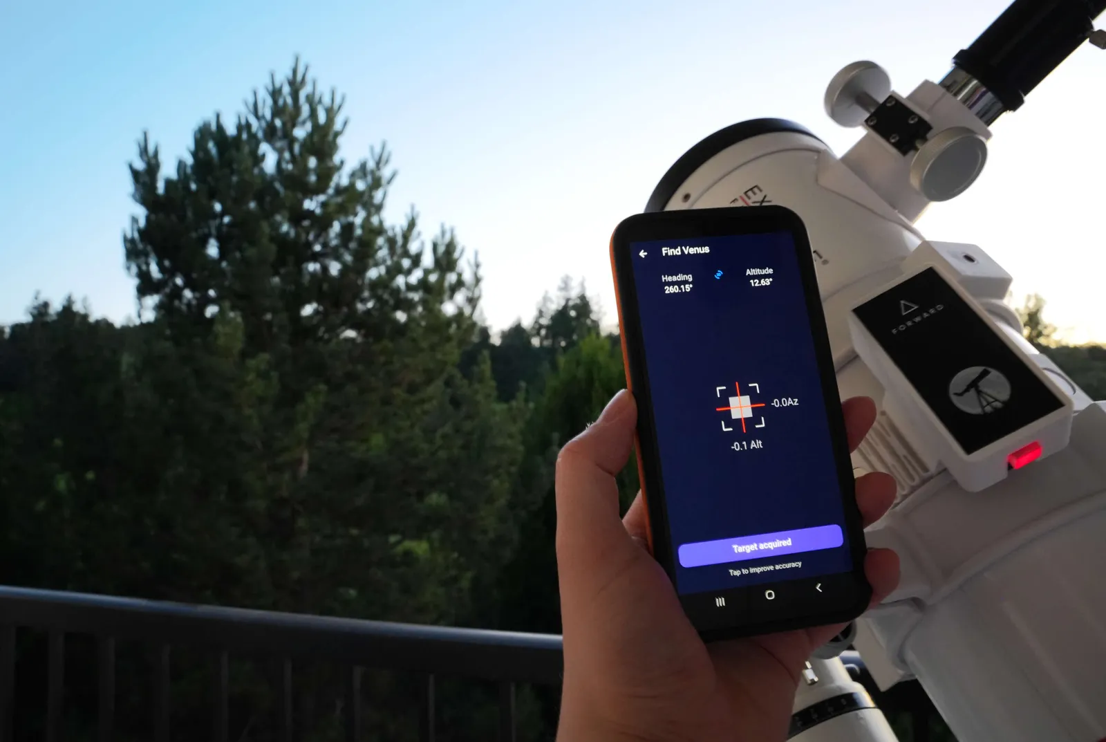 Someone holds a phone next to their telescope, showing the Stargraph interface with Venus locked in. Attached to the telescope is the Stargraph Push-To device with the power button glowing red.